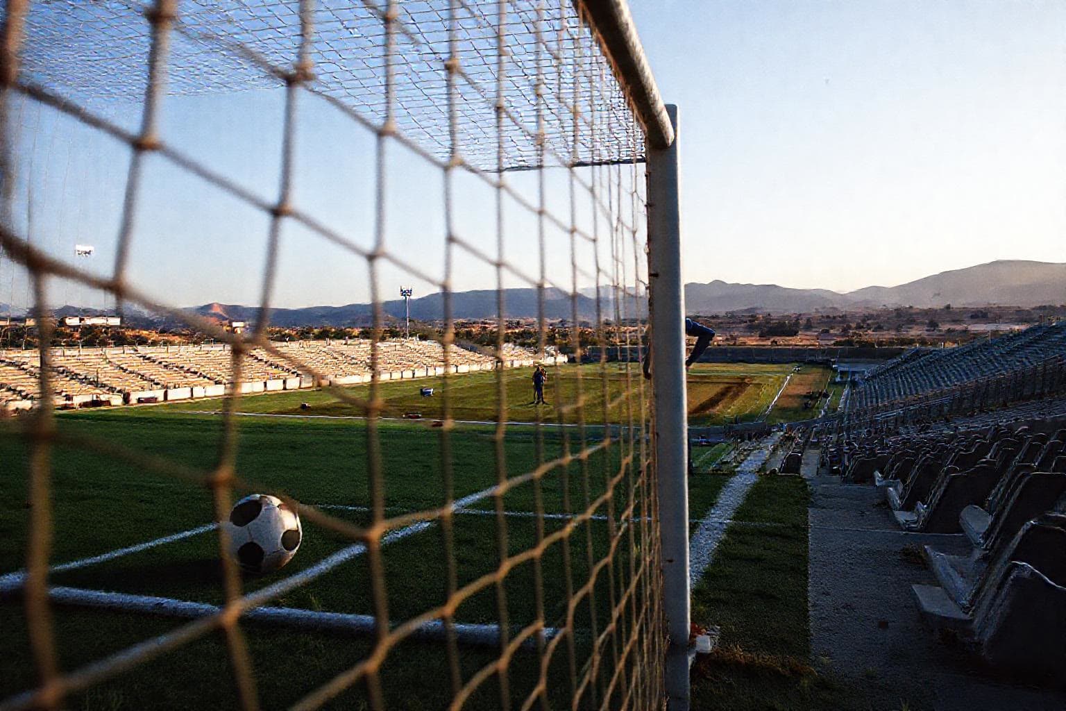 Yeni Karaman Stadyumu'nda kalenin ağında duran futbol topu; arka planda boş tribünler ve arka planda sırtları dönük, yüzleri görünmeyen birkaç kişi; akşamüstü ışığı
