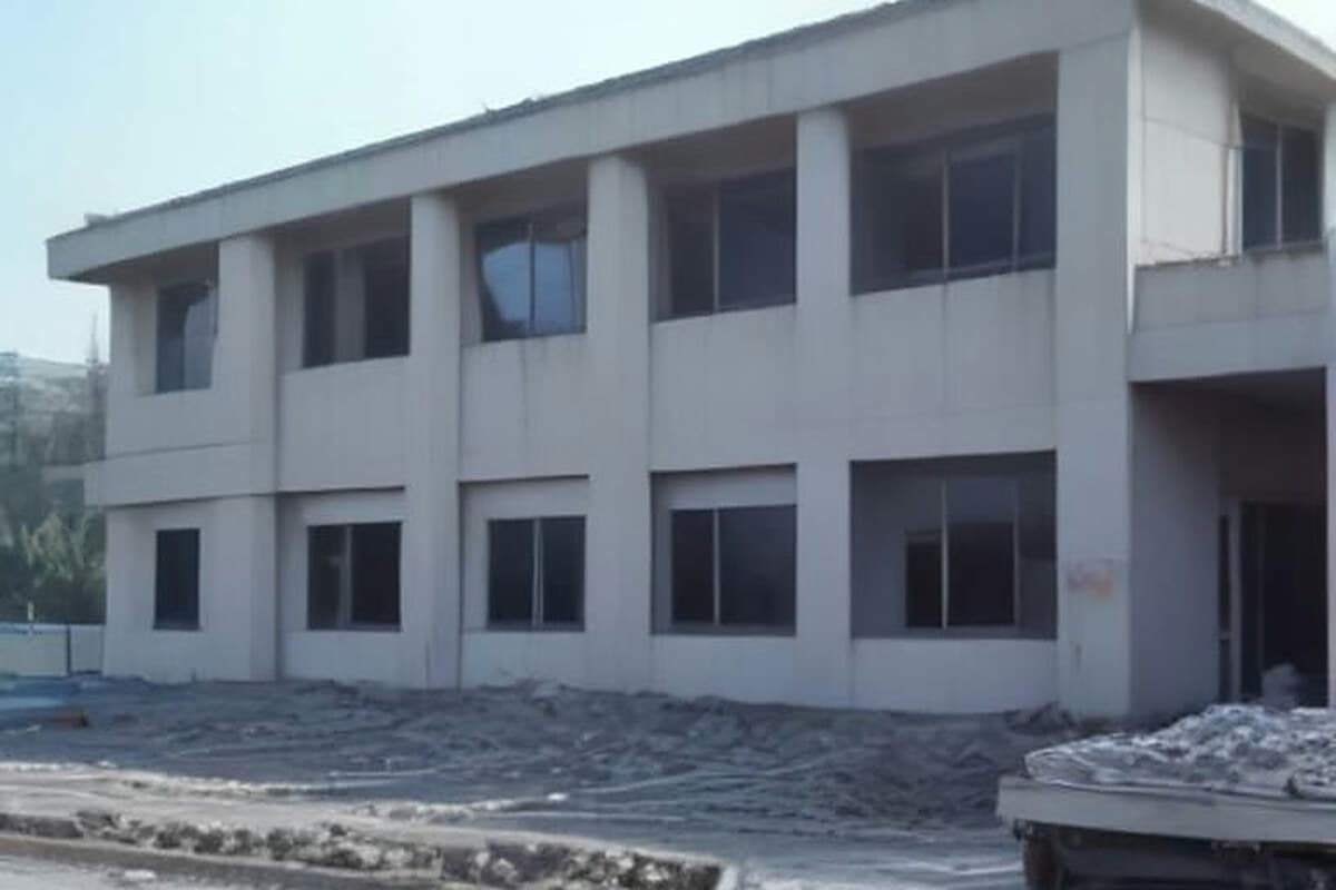 Yeni Sanayi Sitesi içindeki 1987 yapımı Mesleki Eğitim Merkezi binasının önünde akşamüstü yıkım hazırlıkları: bina girişine çekilmiş geçici güvenlik çiti, çevrede dolu kamyon ve küçük bir ekskavatör, uzak planda iş elbiseli, kimliksiz iki kişi (arkadan) çalışıyor; pencere ve kapılar kapatılmış, eğitim binası boş görünümünde.