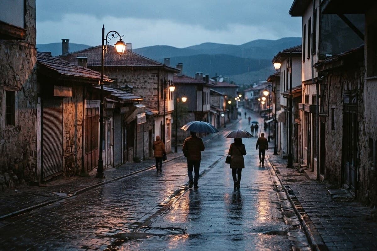 Karaman merkezine benzeyen bir Anadolu kasaba sokağında, akşamüstü çöken yağış bulutları altında ıslak kaldırım ve sokak lambalarının yansıması; birkaç kişi uzaktan, küçük ve anonim şekilde şemsiye açıyor; uzakta alçak tepeler veya Torosların silueti görünüyor.